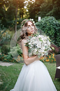 A cute curly woman in a white wedding dress with a wedding bouquet and wreath in her hair standing back to the camera in nature.