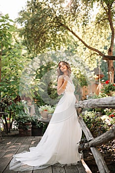 A cute curly woman in a white wedding dress with a wedding bouquet and wreath in her hair standing back to the camera in nature.