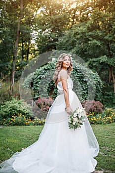 A cute curly woman in a white wedding dress with a wedding bouquet and wreath in her hair standing back to the camera in nature.