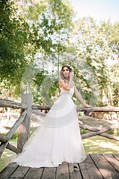 A cute curly woman in a white wedding dress with a wedding bouquet and wreath in her hair standing back to the camera in nature.