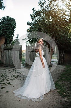 A cute curly woman in a white wedding dress with a wedding bouquet and wreath in her hair standing back to the camera in nature.