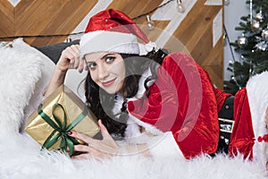 Cute cute girl in clothes santaclaus holding a gift with a green ribbon lying on the bed against the Christmas tree. New year and