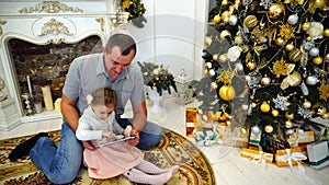 Cute Daddy Daughter And Dad Playing on Tablet Sitting on Floor in Bright Room on Background of Festively Decorated
