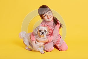 Cute dark haired girl playing with pet dog, looking directly at camera with smile, isolated over yellow background, charming