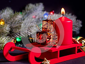 Cute decorative christmas presents on a red sledge with red candle on a backdrop with a fir branch with a christmas garlands