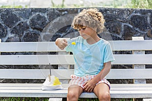 Cute diverse little boy eating a snow cone or shave ice outdoors on a public street bench