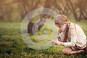 Cute dreamy kid girl in beige outfit picking flowers in spring garden