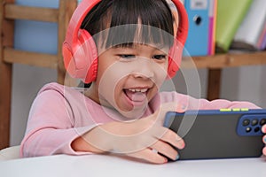 Cute elementary school girl wearing headphones holding a smartphone. Happy Asian kid studying online on smartphone or