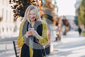 Cute emotional attractive blonde woman in coat with smartphone walks the city street. communication during the walk, lifestyle,