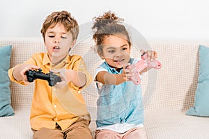 Cute emotional multiethnic kids sitting on couch and playing video game