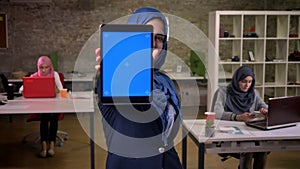 Cute enjoyable female in blue hijab is showing blue screen and holding tablet just at the camera, standing in brick