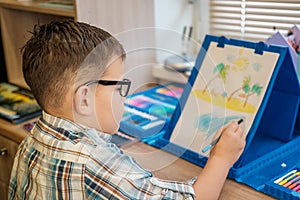 Cute European boy with glasses draws on paper with pencils, sitting at the table