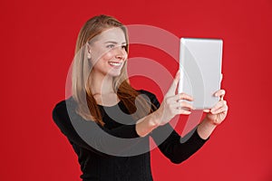 A cute European girl doing selfie on a tablet and smiling. On a red background.