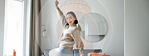 Cute excited asian woman dancing, raising hands up in triumph, celebrating and singing, standing at home in warm jamper