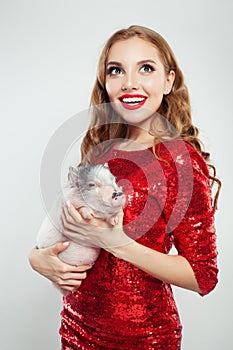 Cute excited woman in red fashionable dress holding mini pig on white background