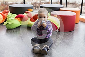 Cute fair baby girl sitting on floor in playroom sucking on toy pensively