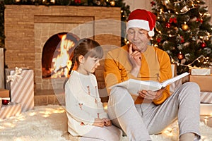 Cute family, grandfather and granddaughter reading book together sitting in festive living room near fireplace and Christmas tree