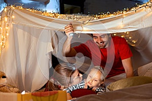 Cute family, mother, father and little daughter lying inside self-made hut, tent in room in the evening and playing