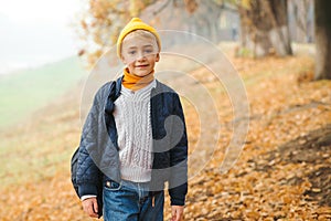 Cute fashionable boy on a walk in autumn park. Stylish kid wears trendy jacket, sweater, jeans and yellow hat. Autumn fashion,