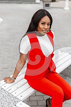 Cute fashionable young black woman in beautiful stylish red suit is resting on wooden bench in city. Attractive luxury trendy