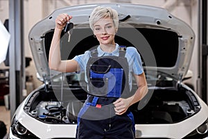 Cute female auto mechanic holding car key at auto repair shop, in overalls uniform