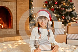 Cute female child in red hat on floor at home. Christmas celebration with presents, little girl posing with gift boxes, looks