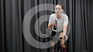 Cute female drinking water or isotonic drink while cycling indoors on cycle smart trainer. Bike pedaling at home workout fitness.