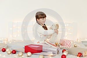 Cute female kid with pigtails wearing white sweater holding soft toy dog sitting on bed with Christmas decoration and garland,