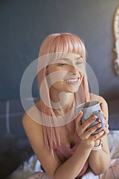 Cute female model holds cup of delicious drink sitting on bed