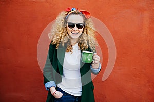 Cute female student with curly blonde hair wearing headband, sunglasses and stylish jacket holding cup of coffee looking at camera