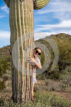 Cute female woman stands next to a large Saguaro cactus in the Sonoran desert, giving it a hug with her arms