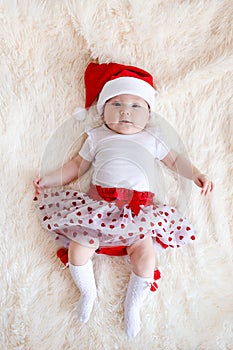 Cute five-month-old baby dressed in a santa hat and a red yuke lies on her back on a fluffy plaid. vertical photo