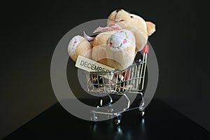 Cute fluffy teddy bear and December word written on wooden stick with coins on mini trolley on dark black background
