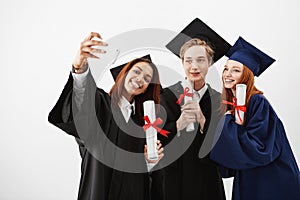 Cute friends graduates smiling holding diplomas making selfie over white background.