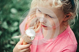 Cute funny adorable girl with dirty nose and milk moustaches eating licking ice cream from waffle cone. Child eating tasty sweet