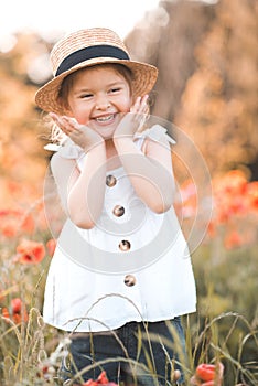 Cute funny baby girl 2-3 year old wear straw hat and white summer dress over flower meadow background. Kid laughing outdoors over