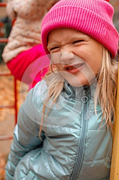 Cute funny blond preschool girl making faces outdoors on a autumn day. Girl wearing pink beanie and blue wind jacket.