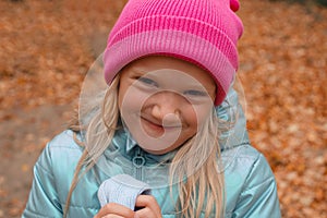 Cute funny blond preschool girl making faces outdoors on a autumn day. Girl wearing pink beanie and blue wind jacket.