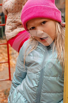 Cute funny blond preschool girl making faces outdoors on a autumn day. Girl wearing pink beanie and blue wind jacket.