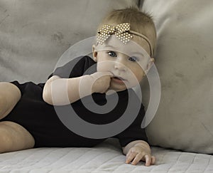 Cute funny caucasian blonde baby girl with brown bow,lying on sofa, looking at camera, putting fingers in mouth,looking