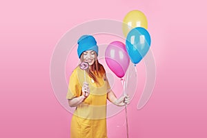 Cute funny girl in blue cap portrait holds an air colorful balloons and lollipop smiling on pink background. Beautiful