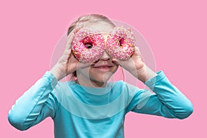 A cute funny girl in a blue t-shirt holds bright pink donuts near her eyes like glasses and looks at you on a pink