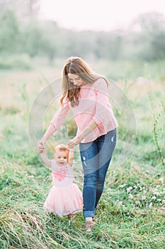 Cute funny happy baby girl making his first steps on a green grass, mother holding her hands supporting by learning to