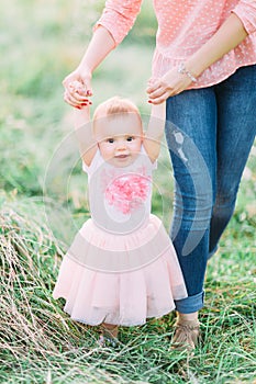 Cute funny happy baby girl making his first steps on a green grass, mother holding her hands supporting by learning to