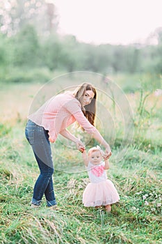 Cute funny happy baby girl making his first steps on a green grass, mother holding her hands supporting by learning to