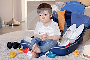 Cute funny little baby boy having rest siiting in blue suitcase tired of packing clothes and toys for vacation.