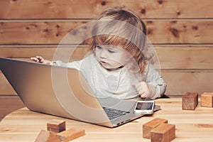 Cute funny little baby boy playing on computer and mobile phone near toy building blocks indoor on wooden background.