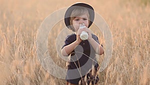 Cute and funny little boy 2-3 years old in a straw hat drinks milk from a glass bottle in a wheat field on a summer day