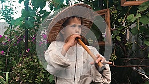 Cute funny little boy in cotton village clothes and straw hat playing on wooden tin wistle. Male child trying to play on flute