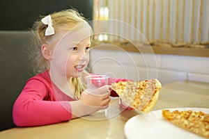 Cute funny little girl eating a slice of pizza at indoor restaurant or cafe